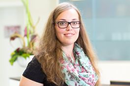 Person mit braunem, langem Haar, Brille, schwarz gekleidet, blumiger Schal.