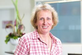 Frau mit blonder, lockiger Frisur, lächelnd; rot weiß kariertes Hemd; unscharfer Hintergrund.