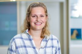 Person mit blonden, lockigen Haaren, lächelt in die Kamera. Trägt kariertes Hemd. Verschwommener Hintergrund.