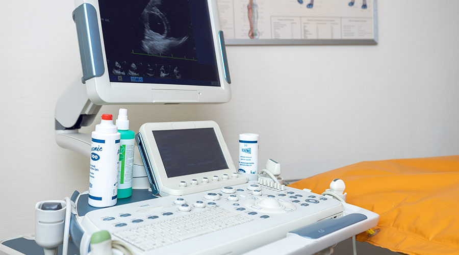 Ultraschallgerät; Monitor mit Bild; Konsole mit Bedienfeld; verschiedene Gelflaschen; gelbe Liege rechts; Wandposter im Hintergrund.