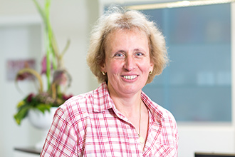 Frau mit blonder, lockiger Frisur, lächelnd; rot weiß kariertes Hemd; unscharfer Hintergrund.