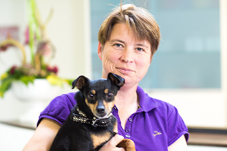 Person in lila Hemd, hält kleinen schwarz-braunen Hund mit gepunktetem Halsband. Neutraler Gesichtsausdruck.