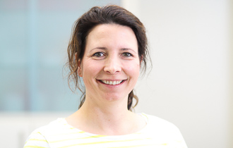 Frau, lächelnd, braune Haare, weiß-gelb gestreiftes Oberteil, neutraler Hintergrund.